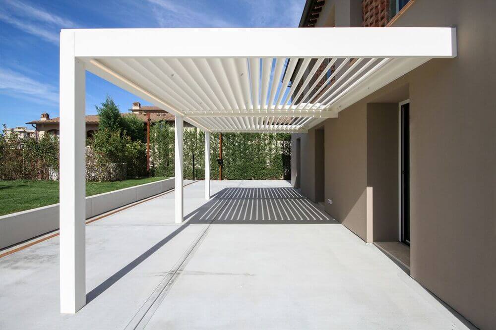 Pergolato bianco con lamelle orientabili, che proietta ombre striate su un patio in cemento.