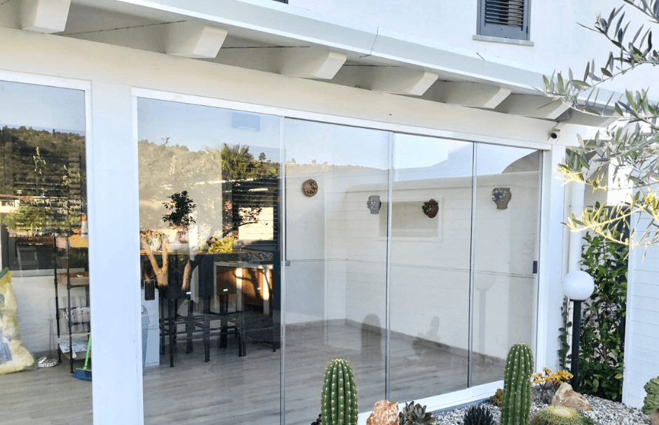 Pergola bianca con vetrate, interno visibile con tavolo e sedie. Esterno con piante e cactus.