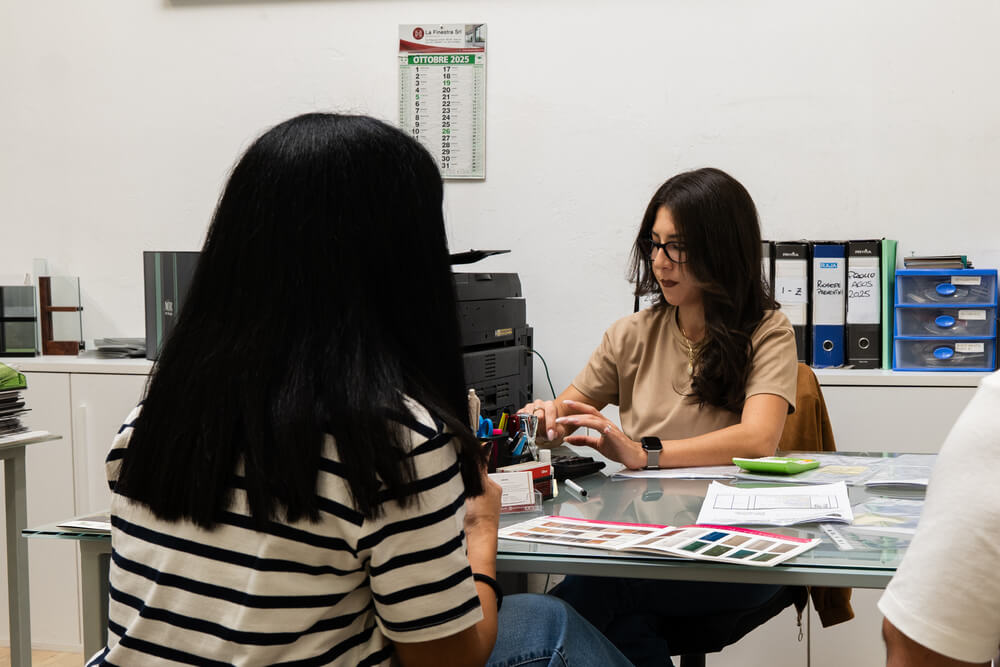 Due donne parlano a un tavolo da ufficio, una ha le spalle rivolte alla telecamera.
