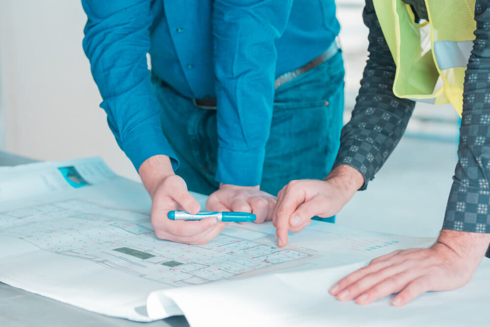 Due uomini esaminano planimetrie edilizie con un pennarello e un dito che indica.