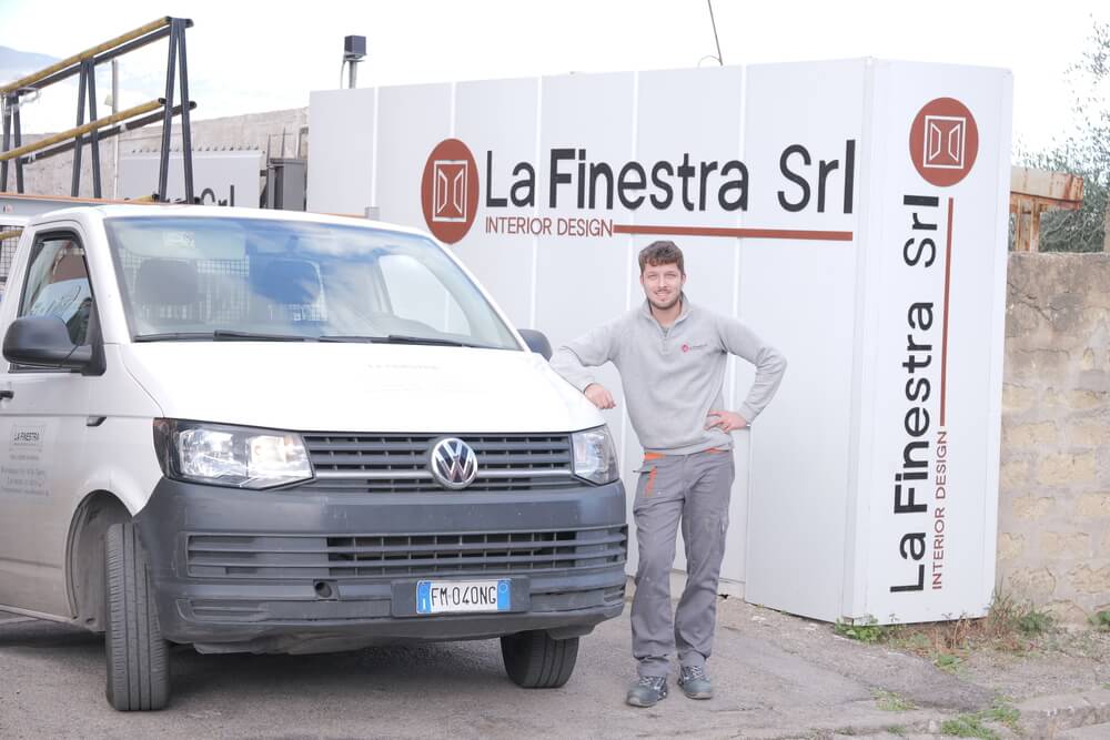 Un uomo sorridente si appoggia a un furgone bianco davanti all'insegna "La Finestra Srl Interior Design".