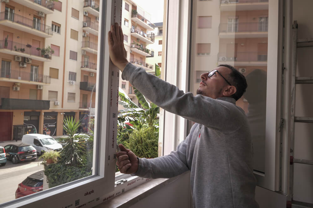 Uomo occhialuto che installa un serramento di finestra, sullo sfondo città.