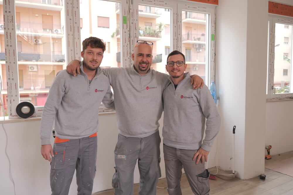 Tre uomini sorridono in abbigliamento da lavoro grigio in una stanza con finestre nuove.